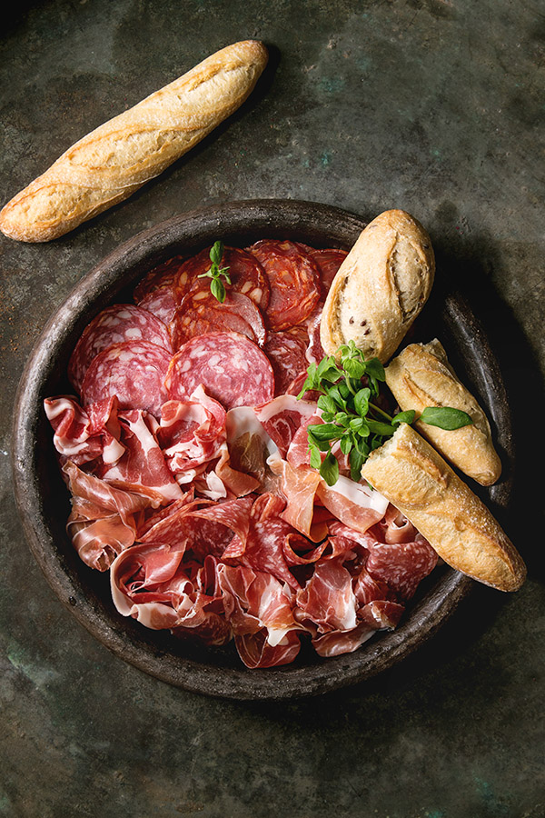Antipasto meat platter assorti of sliced jamon, salami, chorizo sausage in terracotta tray with bread and greens over dark metal background. Flat lay, space